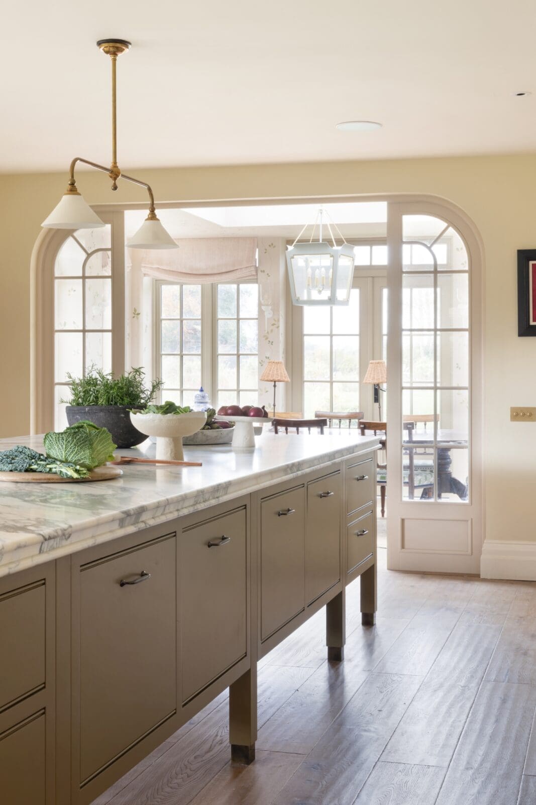 Bright kitchen with a marble island, taupe cabinets, and large arched windows; potted plants and a decorative hanging light create an airy scene.
